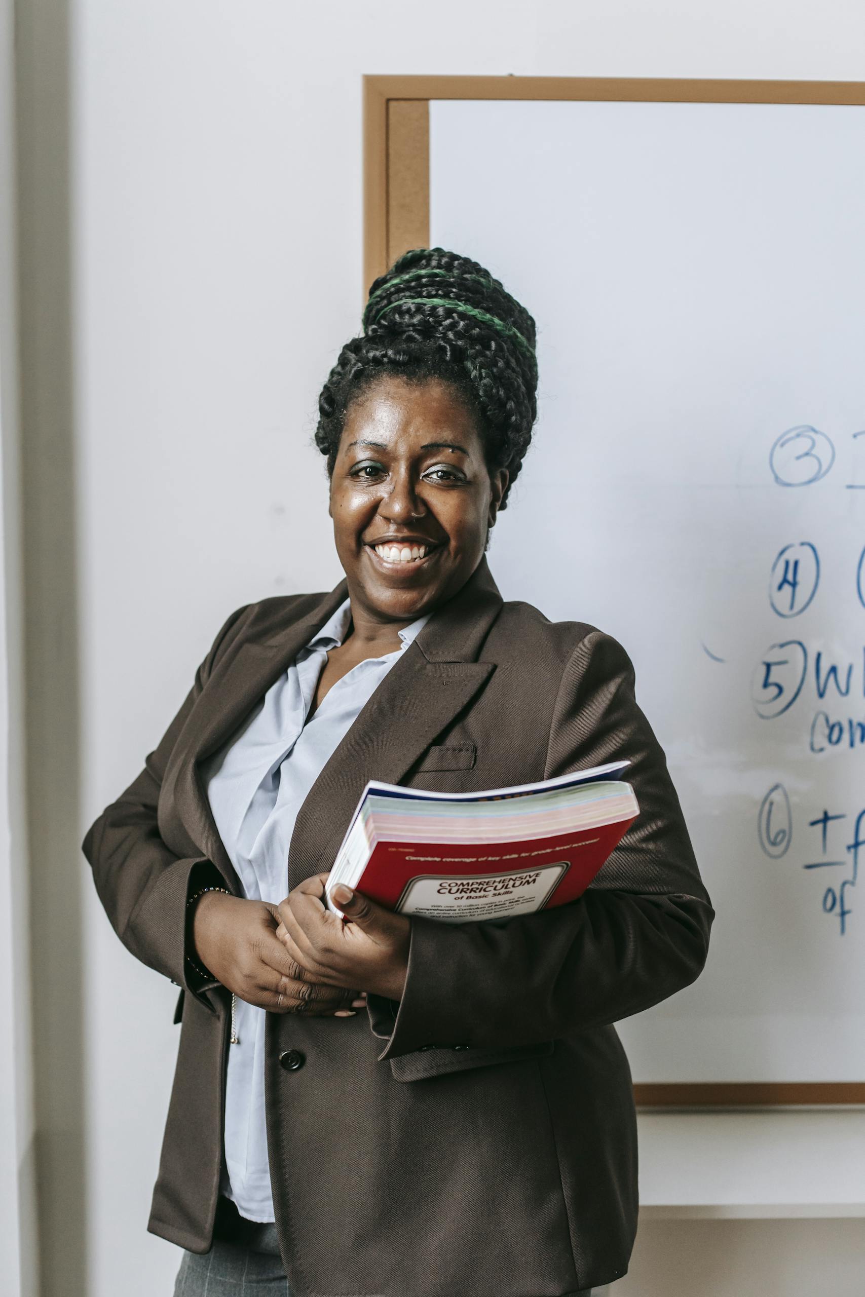 Smiling female leader holding books in front of classroom whiteboard.